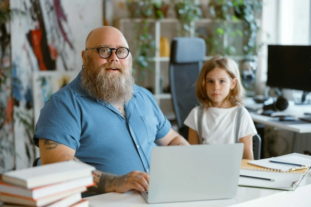 Positive man with glasses helps little daughter to do homework on laptop at table